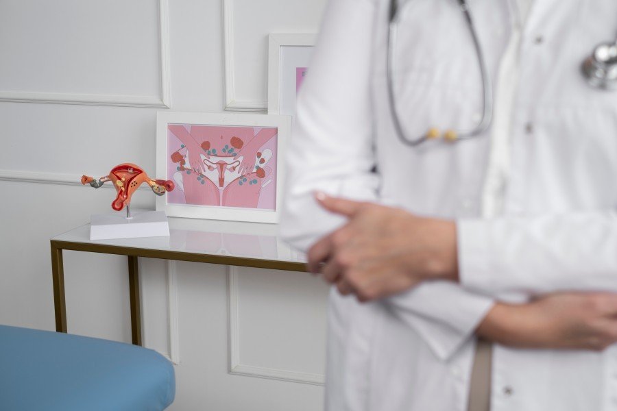 Gynecologist standing in a clinic with a uterus model and female reproductive system illustration in the background.